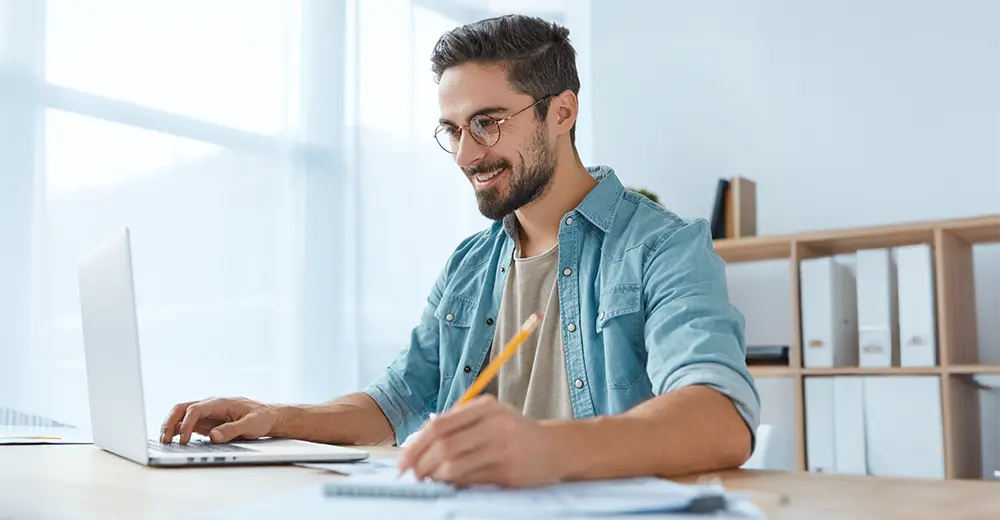 A student sitting at a desk, researching how to file taxes on a laptop and taking notes.