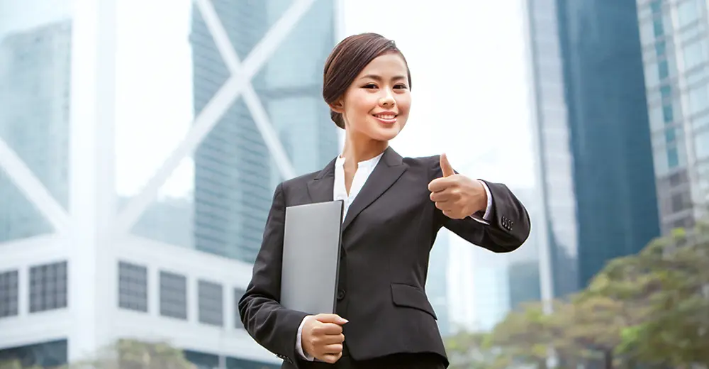 A newcomer to Canada, dressed in business attire and holding a folder, giving a thumbs up in a downtown city setting, ready for work.